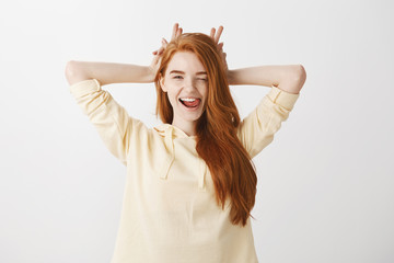 Lifestyle and beautiful people concept. Indoor shot of expressive stylish woman with red hair winking flirty and holding fingers behind head like bunny ears or horns, showing tongue while flirting