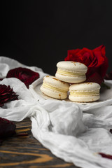 Obraz na płótnie Canvas Three macaroons pastry lying on a plate surrounded by rose blossoms and rose petals on a wooden table covered with a crumpled white cloth