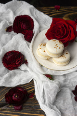 Three macaroons pastry lying on a plate surrounded by rose blossoms and rose petals on a wooden table covered with a crumpled white cloth