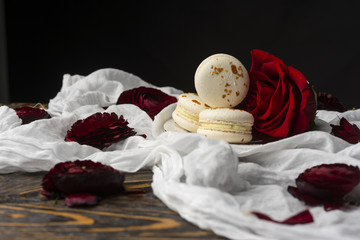 Three macaroons pastry lying on a plate surrounded by rose blossoms and rose petals on a wooden table covered with a crumpled white cloth