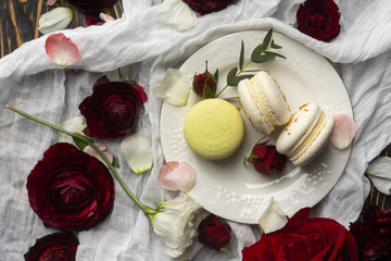 Three macaroons pastry lying on a plate surrounded by rose blossoms and rose petals on a wooden table covered with a crumpled white cloth
