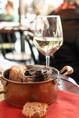 Italian aperitif a glass of white wine and dark olives with pepper and bread, snacks are served in a copper pan on a wooden table.