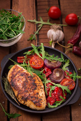 Grilled chicken breast with green salad from arugula and vegetables on a black pan. Wooden rustic background. Top view