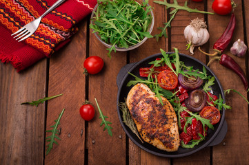 Grilled chicken breast with green salad from arugula and vegetables on a black pan. Wooden rustic background. Top view