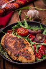 Grilled chicken breast with green salad from arugula and vegetables on a black pan. Wooden rustic background. Top view