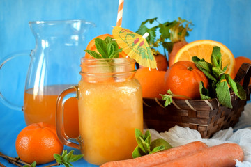 Orange and carrot juice in a glass jar decorated with mint, cocktail straw and umbrella on blue background