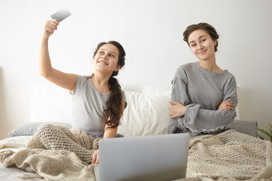 Picture of teenage girl and her mother or elderly sister sitting on bed in front of open laptop, shopping online, ordering food delivery or buying new clothes, browsing web stores using credit card