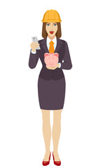 Businesswoman in construction helmet using a mobile phone and holding a piggy bank