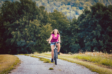 Girl on bicycle cycling down a dirt path in summer, woods in the background