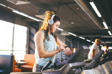 Beautiful young woman with long hair trains in the gym on a treadmill. Listens to music in large headphones. On his arm a sports watch in black for tracking speed, heart rate, pace and stopwatch