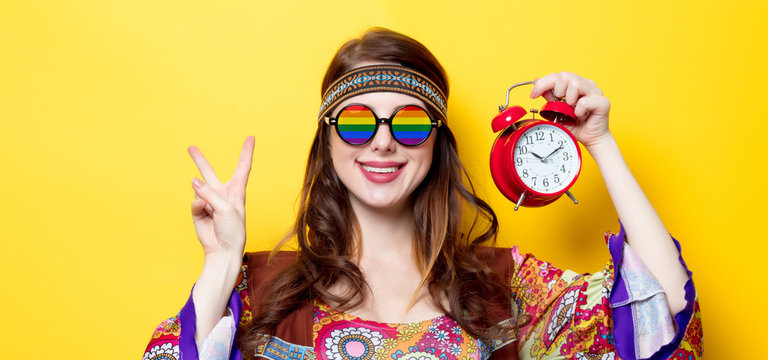 Young hippie girl with rainbow glasses and alarm clock on yellow background