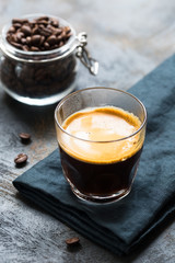 Glass cup of fresh coffee time with thick golden foam froth on dark rustic background, espresso americano on black tissue with spoon of brown sugar and coffee beans, vertical copy space