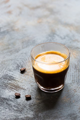 Glass cup of fresh coffee time with thick golden foam froth on dark rustic background, espresso americano on black tissue with spoon of brown sugar and coffee beans, vertical copy space