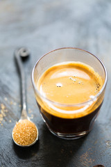 Glass cup of fresh coffee time with thick golden foam froth on dark rustic background, espresso americano on black tissue with spoon of brown sugar and coffee beans, vertical copy space