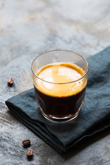 Glass cup of fresh coffee with thick golden foam froth on dark rustic background, espresso americano on black tissue with spoon of brown sugar and coffee beans, vertical copy space