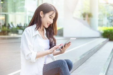 Young and beautiful Asian woman using her tablet PC while sitting on the steps outside the modern office building in the urban city