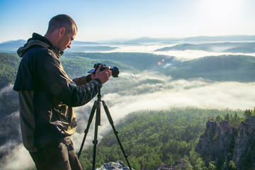 Photographer takes photos with camera on tripod on rocky mountain peak. Beautiful misty sunrise and valley view over clouds. - obrazy, fototapety, plakaty