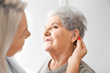 Otolaryngologist putting hearing aid in senior woman's ear on light background