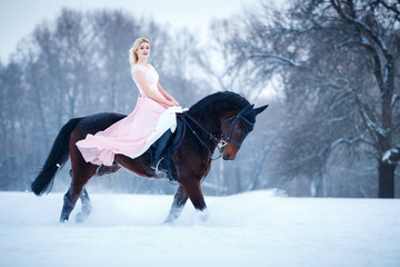 Young woman in rose dress riding her bay horse on winter field. Romantic or historical equestrian...