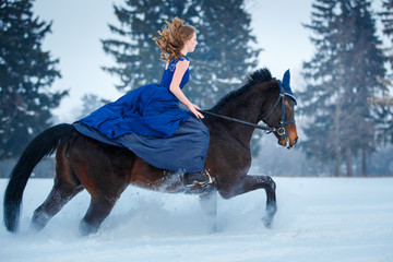 Young girl in blue dress galloping horseback on winter field. Romantic or historical equestrian...