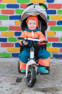Happy boy in bright suit is riding children's bicycle wheelchair against background of multicolored brick wall. Romantic lighting with sunlight reflections. Lens flare effect without post prodaction