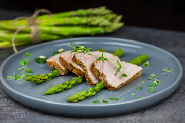 Slices of roasted pork tenderloin on asparagus with pesto from kale and thyme on the grey plate. A bunch of asparagus on the background