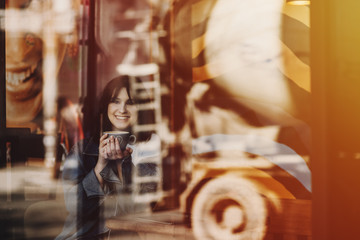 Happy young woman sitting in a coffee shop and holding in hands a cup while is looking at camera through window.