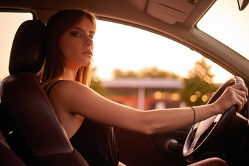 Fototapeta premium Concept: strong independent young woman in car. Beautiful serious girl sit behind the wheel at twilight. Inside look.