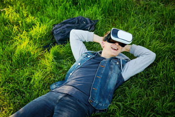 The young man lies on a green grass with VR glasses and relaxing. Outdoor. Copy space.