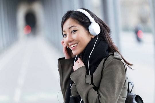 Smiling asian young woman listening to music and looking at camera in the street.