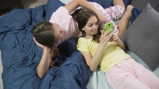 friendly relaxed young teenage girls chatting and browsing smartphone on bed at leisure time. modern digital device and gadgets addiction.