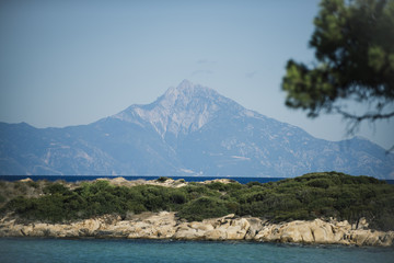 Beautiful landscape with high mountain, covered with snow. Light rocky top of mountain and blue sky, horizon, skyline. Sea and stony seashore landshaft covered with green plants. Nature on sunny day.
