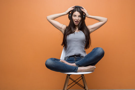 Portrait of a young beautiful girl sitting in the chair and listening to music through the headset, her hands on her head, against orange background. Lifestyle, people and technology concept