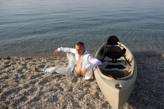 Man sits on beach near canoe boat on sunny summer day, sea surface in background. Macho with torso spend leisure at seashore. Vacation concept. Guy on strict face wears white shirt on vacation, resort