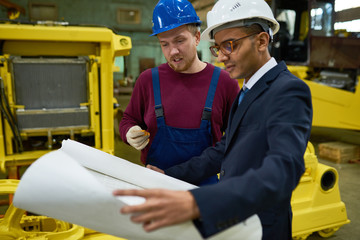 Handsome mixed-race entrepreneur wearing protective helmet and suit studying blueprint with factory worker while standing at spacious production department