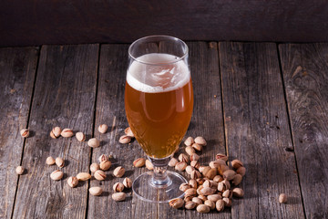 A glass of refreshing light beer and a scattered handful of pistachios on a table of old textured wooden boards.