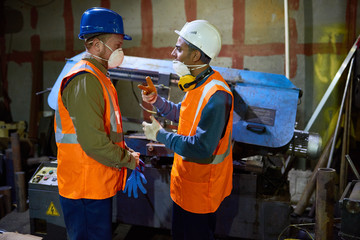 Profile view of machine operators wearing filter masks and reflective vests discussing results of accomplished work while standing at spacious production department of modern plant