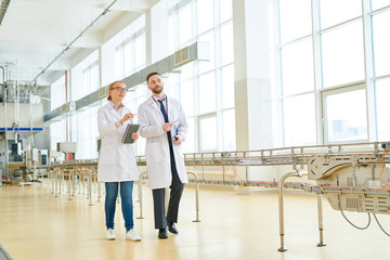 Confident technologists wearing white coats walking along spacious production department of milk factory and carrying out quality control