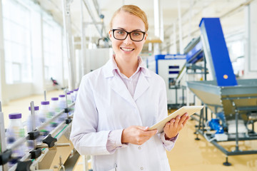 Attractive young inspector wearing white coat looking at camera with charming smile while standing at spacious soy milk factory with digital tablet in hands, waist-up portrait shot
