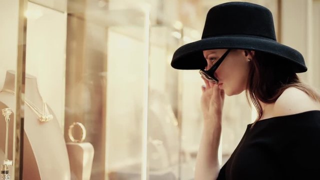 A beautiful stylish rich girl in a hat takes off her glasses to see the goods on the shop window of a luxury boutique. Slow motion