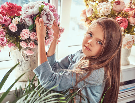 Portrait of a cute little girl with long brown hair and piercing glance wearing a stylish dress, posing with flowers against window at home