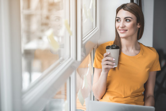Portrait of good looking girl relaxing on windowsill with cup of beverage in hand. She is smiling. Copy space in left side