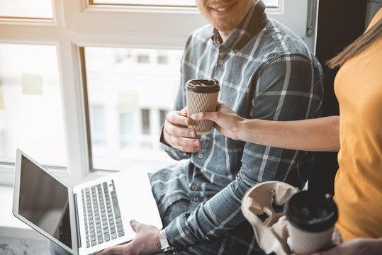 Woman extending hand with drink to male colleague. Man sitting with laptop and taking cup