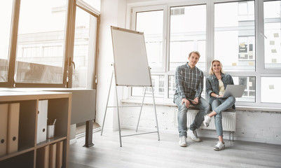 Portrait of man and woman sitting near office window and smiling at camera. They keeping gadgets in hands. Copy space in left side