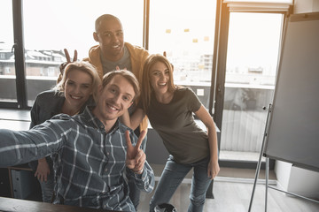 Portrait of four happy people making snapshot of themselves at work. They are showing peace sign taking selfie in office