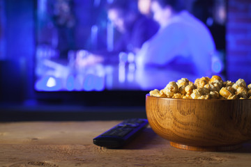 A wooden bowl of popcorn and remote control in the background the TV works. Evening cozy watching a movie or TV series at home