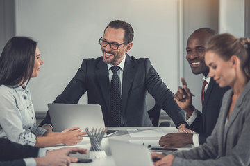 Outgoing bearded employer talking with cheerful colleagues while sitting at table and working with laptop in office. Beaming partners during labor concept