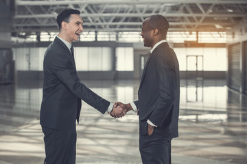 Side view laughing employer shaking hand of beaming male while standing in big empty buildng. Happy partners during agreement concept