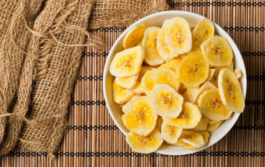 Heap of dried banana chips snack in white bowl on bamboo mat