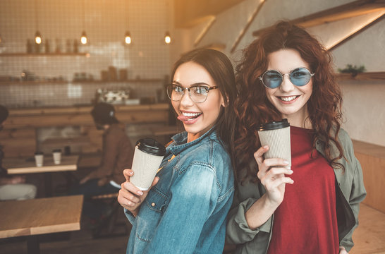Waist up portrait of two outgoing girls standing and posing in cafeteria. They are holding cups of espresso and laughing. Pleasure and fun concept